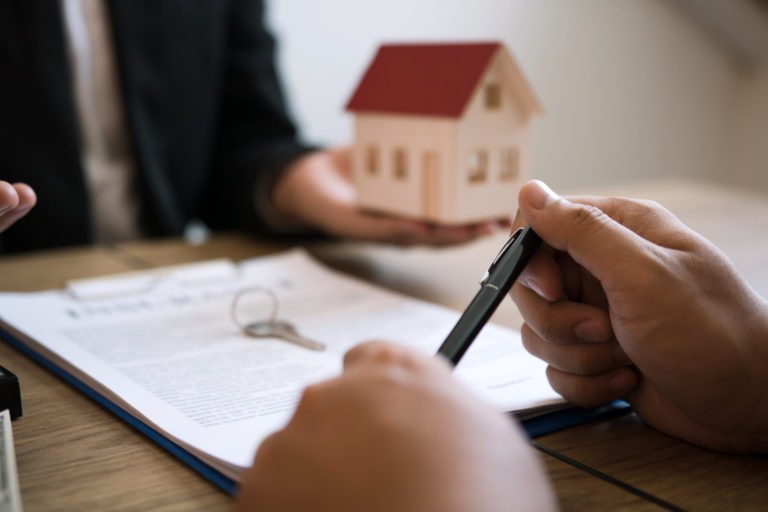 a person holding a pen over a paper with a house in the background